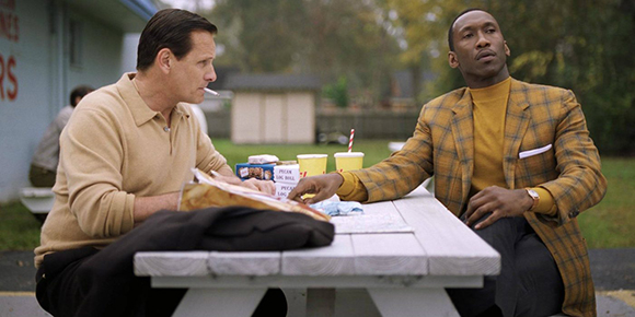 Two men sit at a white wooden picnic table outdoors with drinks and food containers.