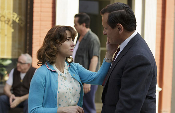 A woman in a blue cardigan reaches out to touch the face of a man in a dark suit.