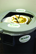 A gray compost bin with a lid containing a white plate of food scraps and a green label.