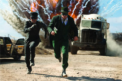 Two men in dark suits run toward the camera as a large explosion erupts behind a truck on a dirt road.