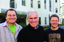 Three men standing side-by-side and smiling in front of a white multi-story building.