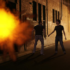 Two men stand in a dark alley near a large, bright orange explosion.