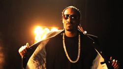 A man wearing sunglasses, a black shirt, and a thick silver chain stands against a dark, backlit background.