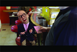 A man with a disproportionately large head sits at a diner counter, pointing at a pink slip.