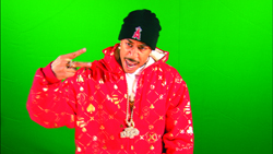 A man wearing a red patterned hoodie and black beanie gestures with his hand against a green background.