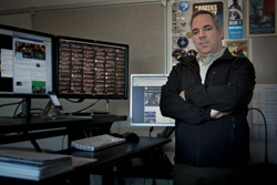 A man with arms crossed standing in front of several computer monitors in an office.