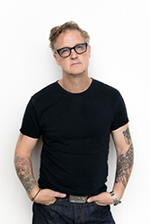 A man with glasses and tattoos on both arms wearing a black t-shirt and dark jeans.