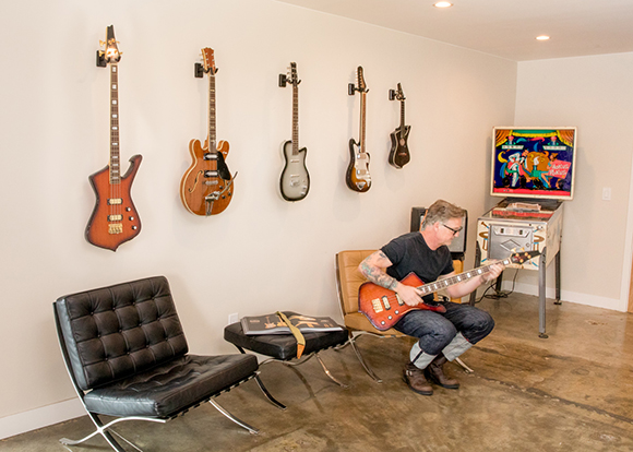 A man sits in a chair playing a red electric guitar in a room with five guitars hanging on the wall.