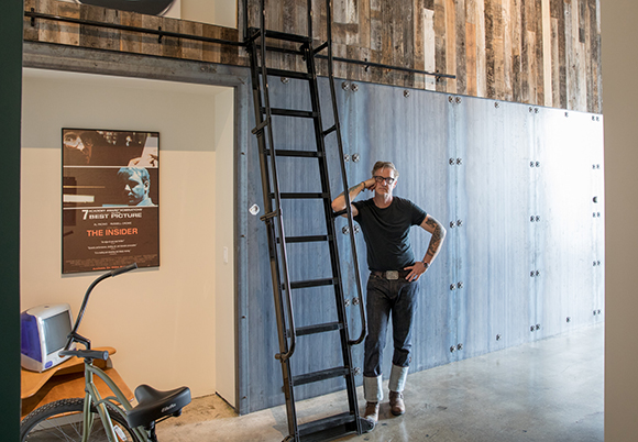 A man in a black t-shirt and jeans stands beside a tall black metal ladder in an industrial-style room.