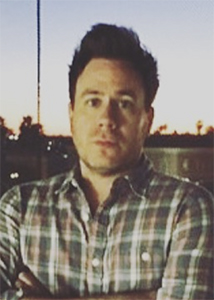 A man with dark hair wearing a plaid button-down shirt, looking forward against a twilight sky.