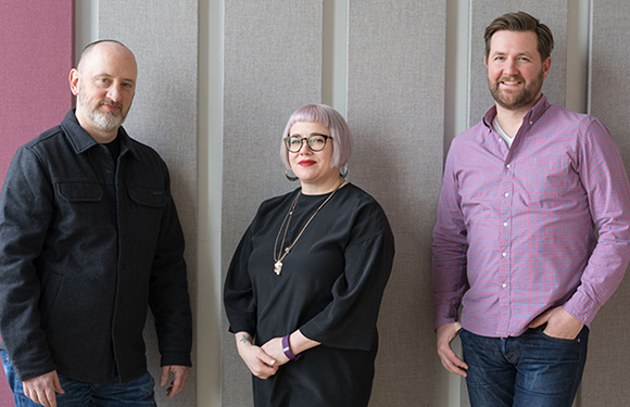 A man in a black jacket, a woman with pink hair in a black top, and a man in a purple shirt stand against a wall.