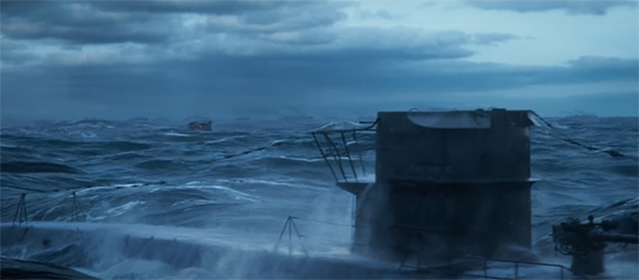 A submarine's conning tower emerges from turbulent, dark blue ocean waves under a heavy, overcast sky.