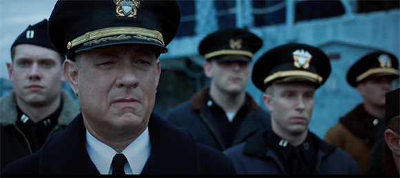 A man in a naval officer's cap looks forward with a serious expression, surrounded by other uniformed men.