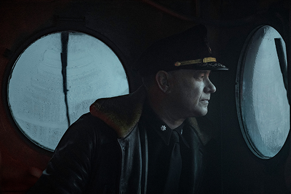 A man in a dark military uniform and peaked cap looks through a circular porthole in a dark setting.