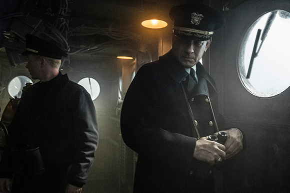 Two men in dark naval uniforms stand inside a dimly lit ship near circular portholes.