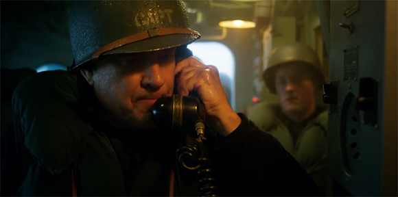 A man wearing a military helmet speaks into a black telephone handset in a dimly lit interior.