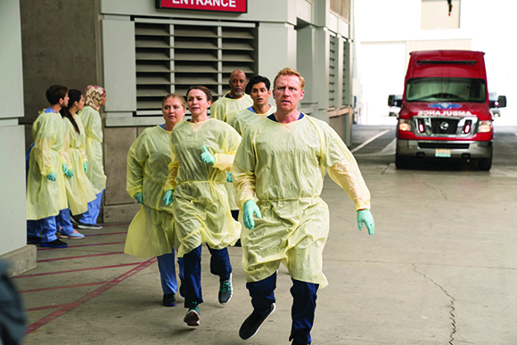 Medical staff in yellow gowns and blue scrubs walk urgently toward the camera in a hospital driveway.