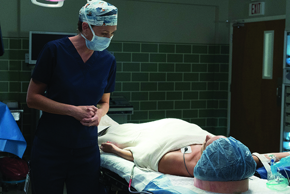 A medical professional in blue scrubs and a surgical mask looks down at a patient lying on an operating table.