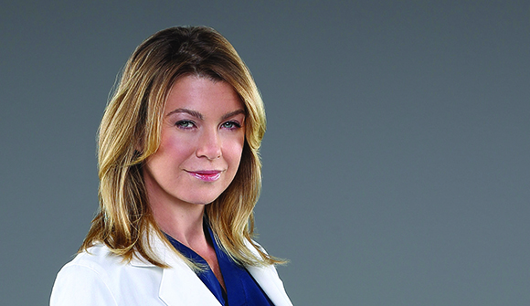 A woman with blonde hair wearing a white lab coat and blue shirt against a gray background.