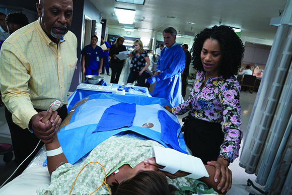 Medical staff and observers stand around a patient lying on a gurney in a hospital room.