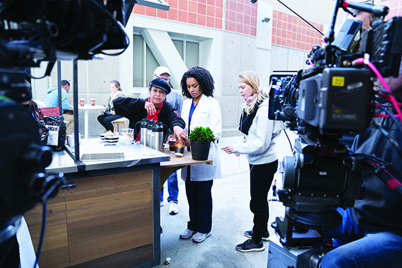 A film crew uses large cameras to film a woman in a white lab coat standing at a table outdoors.