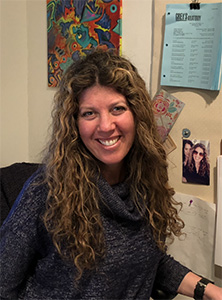 A smiling woman with long, curly brown hair wearing a blue textured sweater.