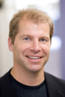 A smiling man with short blond hair wearing a black shirt and jacket.