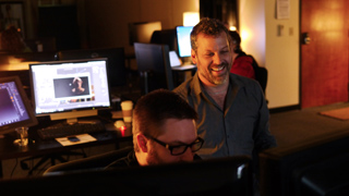 A man smiles while looking at a computer screen next to a seated colleague in a dark office.
