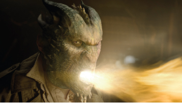 A green, horned reptilian creature exhales a large stream of bright orange fire from its mouth.