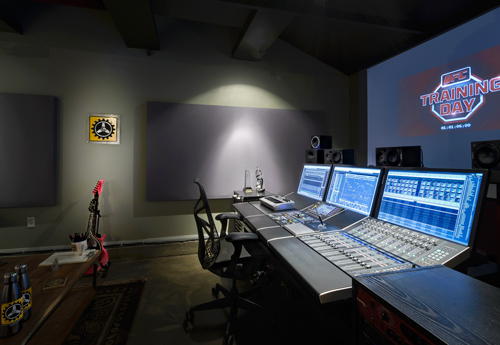 A large digital audio mixing console in a dark studio with a projection screen in the background.