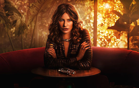 A woman with wavy hair sits on a red sofa with her arms crossed in a warm, golden-lit room.