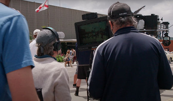 A film crew works outdoors with a large professional camera and monitor on a tripod.
