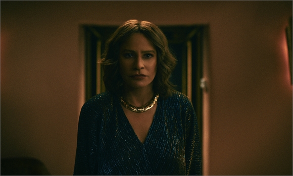 A woman with wavy hair and a gold necklace wearing a dark blue shimmering top looks forward with a serious expression.