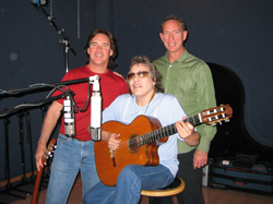 Three men in a recording studio, one seated playing an acoustic guitar and two standing behind him.