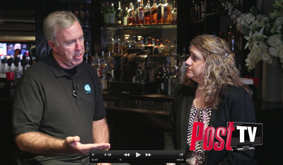 A man in a black polo shirt gestures while talking to a woman with curly hair in a bar setting.