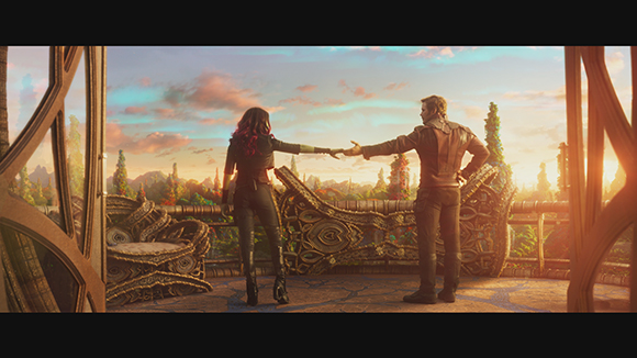 A woman and a man stand on a balcony, reaching for each other's hands against a sunset sky.