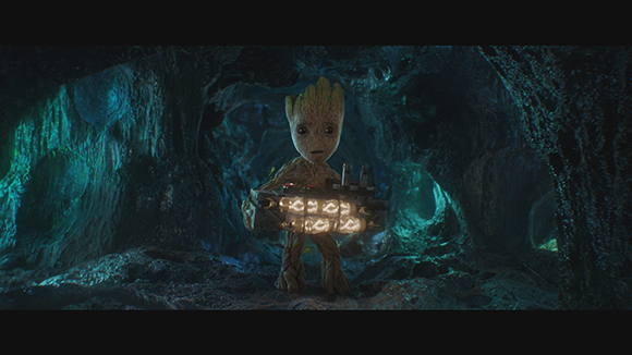 A small tree-like creature holds a glowing, rectangular technological device inside a dark, blue-lit cavern.