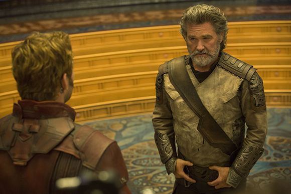 Two men in detailed, earth-toned sci-fi armor stand in a large, circular room with tiered seating.