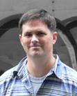 A man with short dark hair wearing a blue and white striped button-down shirt.