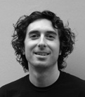 A smiling man with dark, curly hair looking toward the camera in a black and white portrait.