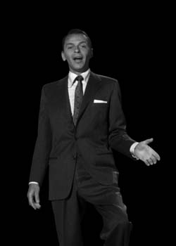 A man in a dark suit and tie singing with his mouth open and one hand extended against a black background.