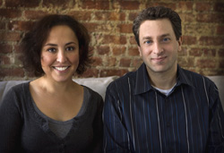 A smiling woman in a dark grey shirt and a man in a blue striped shirt sit in front of a brick wall.