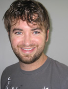 A smiling man with wavy brown hair and a short beard wearing a grey t-shirt.