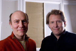 Two men smiling side by side indoors.