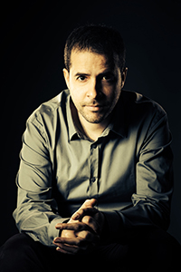 A man with short dark hair and a light beard wearing a grey button-down shirt, looking directly at the camera.