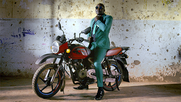 A man in a green suit and sunglasses stands next to a red motorcycle in a weathered room.