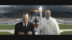 A man in a suit and a man in a white cloak holding a staff stand before statues under a dark sky.