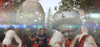 People dancing in a forest under two large, glowing, metallic spherical objects in the sky.