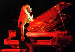 A blonde woman in a black dress sits at a grand piano illuminated by intense red stage lighting.