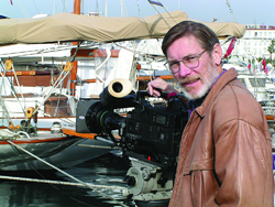 A man in a brown leather jacket stands beside a large professional film camera on a boat.